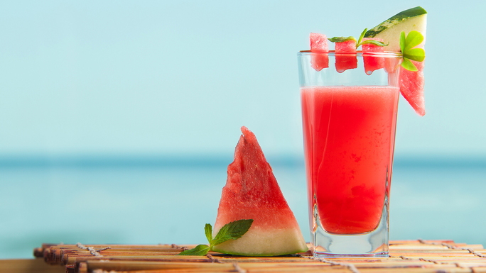 Glas mit Wassermelonen-Smoothie auf Bambus-Untersetzer und Wassermelonenstück als Dekoration, im Hintergrund unscharf Wasseroberfläche