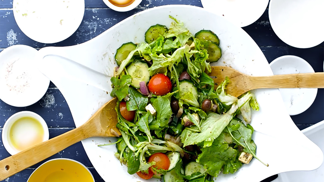 Angerichteter Sommersalat in weißer Schüssel, mit Salatbesteck aus Holz, Zubereitungsschalen im Hintergrund.