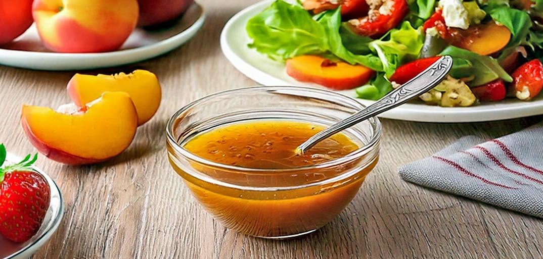 A bowl of homemade dressing placed on a wooden table, accompanied by fresh strawberries, peach slices, and a vibrant salad on a plate, all set in a warm, inviting atmosphere.