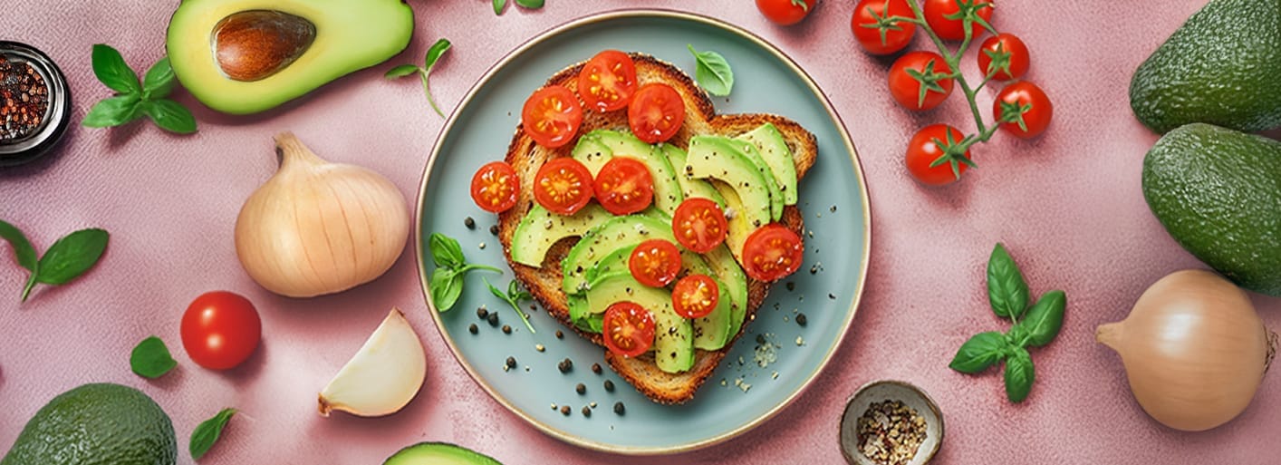 Heavenly Avocado Tomato Toast _Header A plate of avocado toast topped with sliced avocado and cherry tomatoes, surrounded by fresh herbs, an avocado, onion, garlic, and seasoning