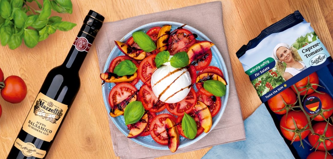 A colorful salad with fresh tomato and grilled peach slices arranged around a ball of burrata cheese, garnished with fresh basil and drizzled with balsamic vinegar. Next to the plate are vine tomatoes, a bottle of Mazzetti Aceto Balsamico, and a pack of SanLucar cocktail tomatoes.