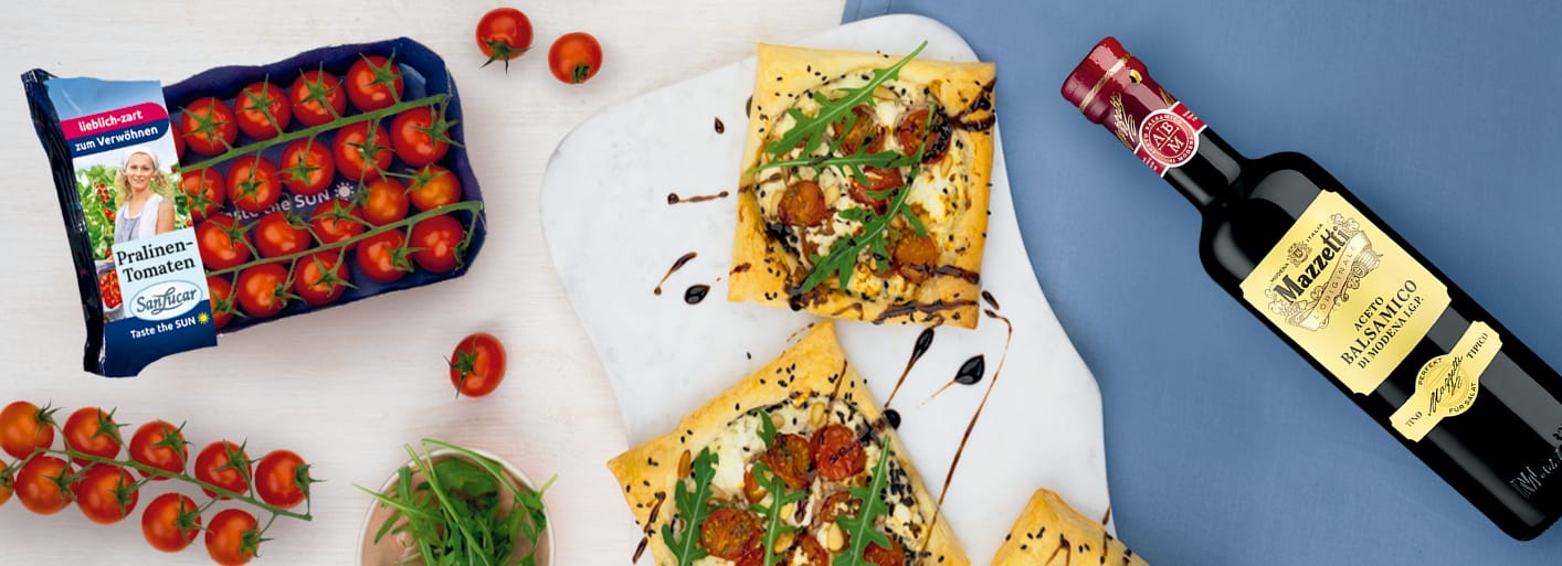Würzige Blätterteigschiffchen mit Tomaten_Header Rectangular puff pastry tarts topped with cherry tomatoes, creamy cheese, fresh arugula, and balsamic glaze on a light serving board. Nearby are a cluster of vine tomatoes, a bottle of Mazzetti balsamic vinegar, and a pack of SanLucar Pralinen-Tomaten tomatoes.